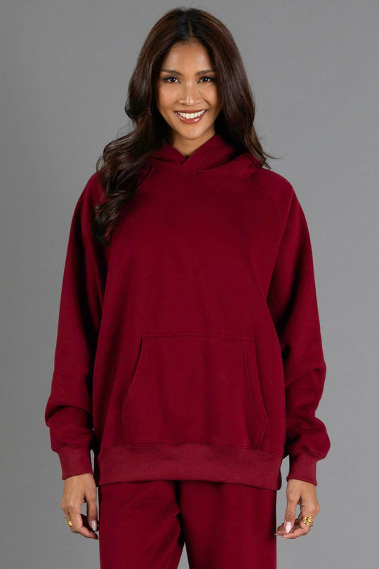 A woman with long dark hair, smiling, wears the Maroon Panache Hooded Sweatshirt by Panache Apparel Collection and matching pants, standing against a plain gray background.
