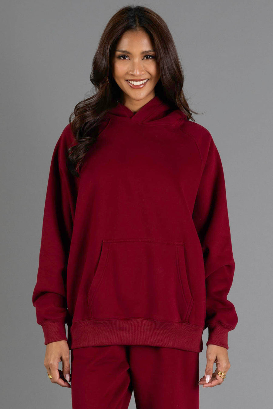 A woman with long dark hair, smiling, wears the Maroon Panache Hooded Sweatshirt by Panache Apparel Collection and matching pants, standing against a plain gray background.