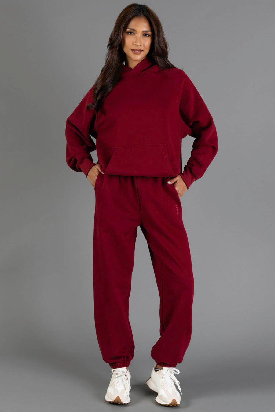 A woman stands against a gray background wearing the Maroon Panache Hooded Sweatshirt and matching sweatpants from the Panache Apparel Collection, paired with white sneakers. She places her hands on her hips, looking at the camera with a soft smile.