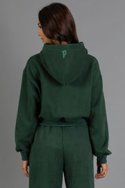 A woman with long brown hair, seen from behind, wears the Panache Apparel Collection’s Dark Green Panache Cropped Hooded Sweatshirt with matching pants. The hoodie features a small light green logo on the hood. She stands against a plain gray background.