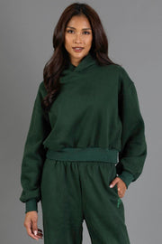 A woman with long dark hair wears the Dark Green Panache Cropped Hooded Sweatshirt from the Panache Apparel Collection and matching pants, standing against a plain gray background, looking at the camera and smiling slightly.