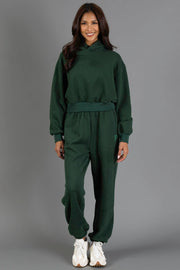 A woman stands smiling in a Dark Green Panache Cropped Hooded Sweatshirt by Panache Apparel Collection, paired with matching jogger pants and white sneakers, against a plain gray background.