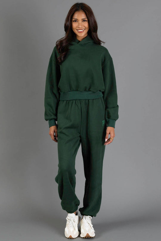 A woman stands smiling in a Dark Green Panache Cropped Hooded Sweatshirt by Panache Apparel Collection, paired with matching jogger pants and white sneakers, against a plain gray background.