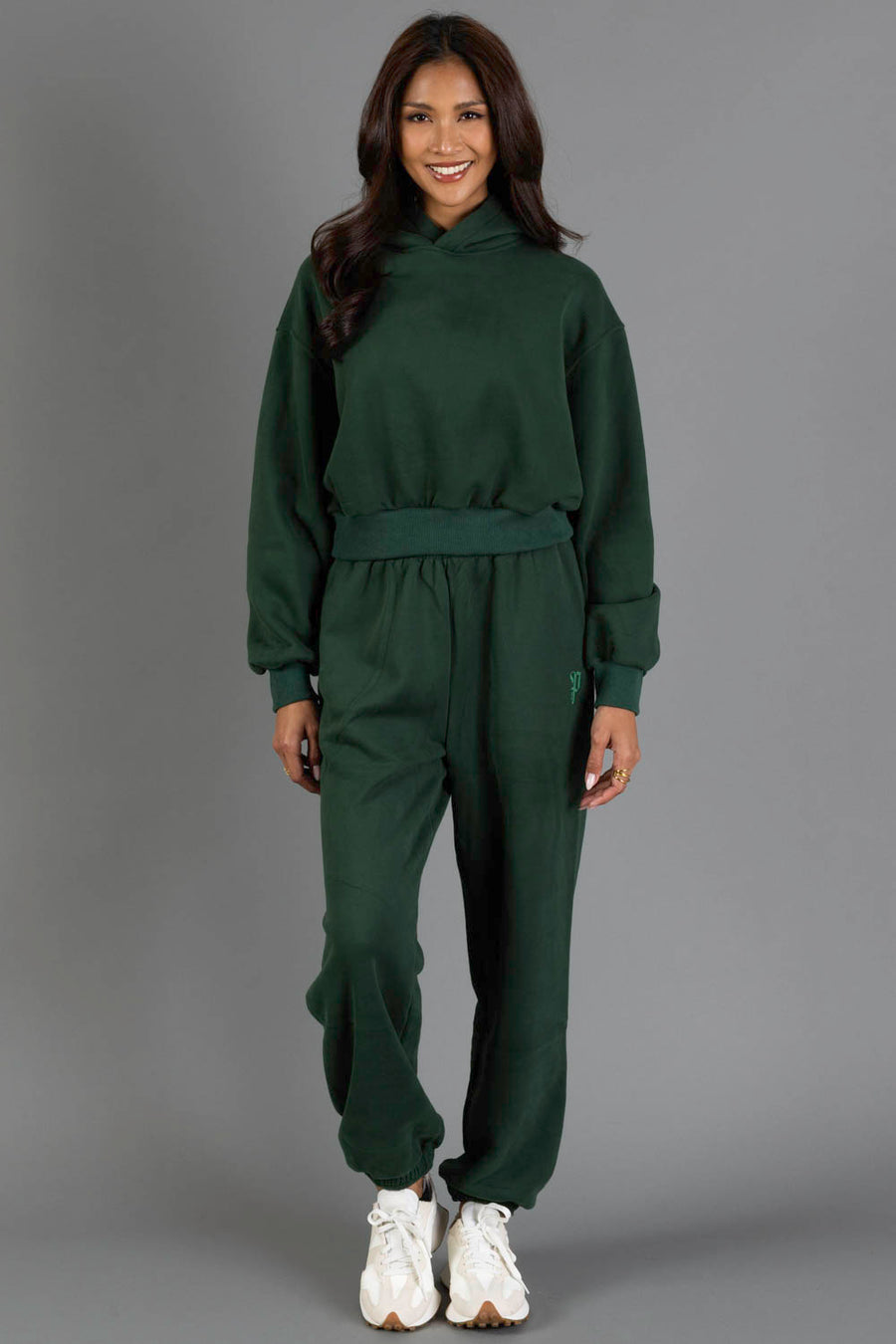 A woman stands against a gray background, smiling at the camera while wearing Panache Apparel Collection’s Dark Green Panache Sweatpants paired with a matching hoodie and white sneakers.