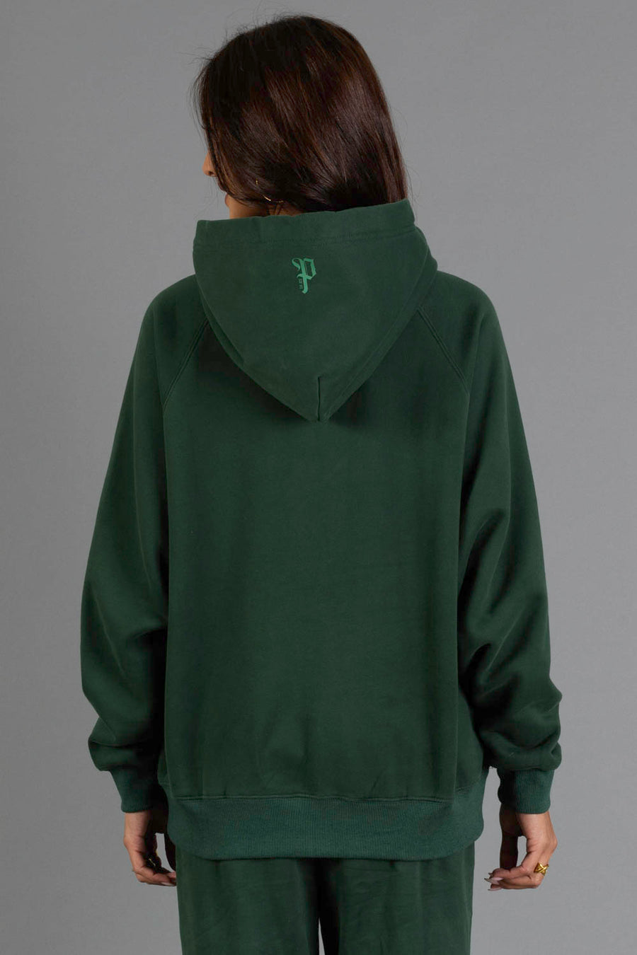 A person with long brown hair, facing away from the camera, is wearing the Panache Apparel Collection’s Dark Green Panache Hooded Sweatshirt and matching pants. The sweatshirt features a small embroidered letter “P” on the back of the hood. The background is plain gray.