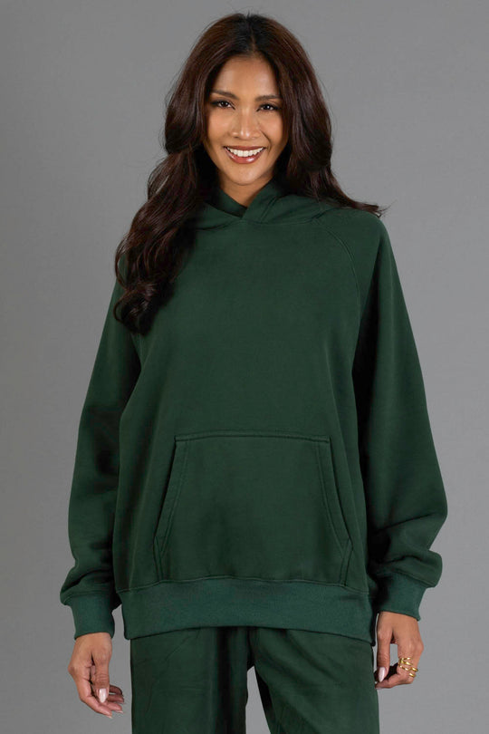 A woman with long dark hair smiles while wearing the Panache Apparel Collection’s Dark Green Panache Hooded Sweatshirt and matching pants, standing against a plain gray background.