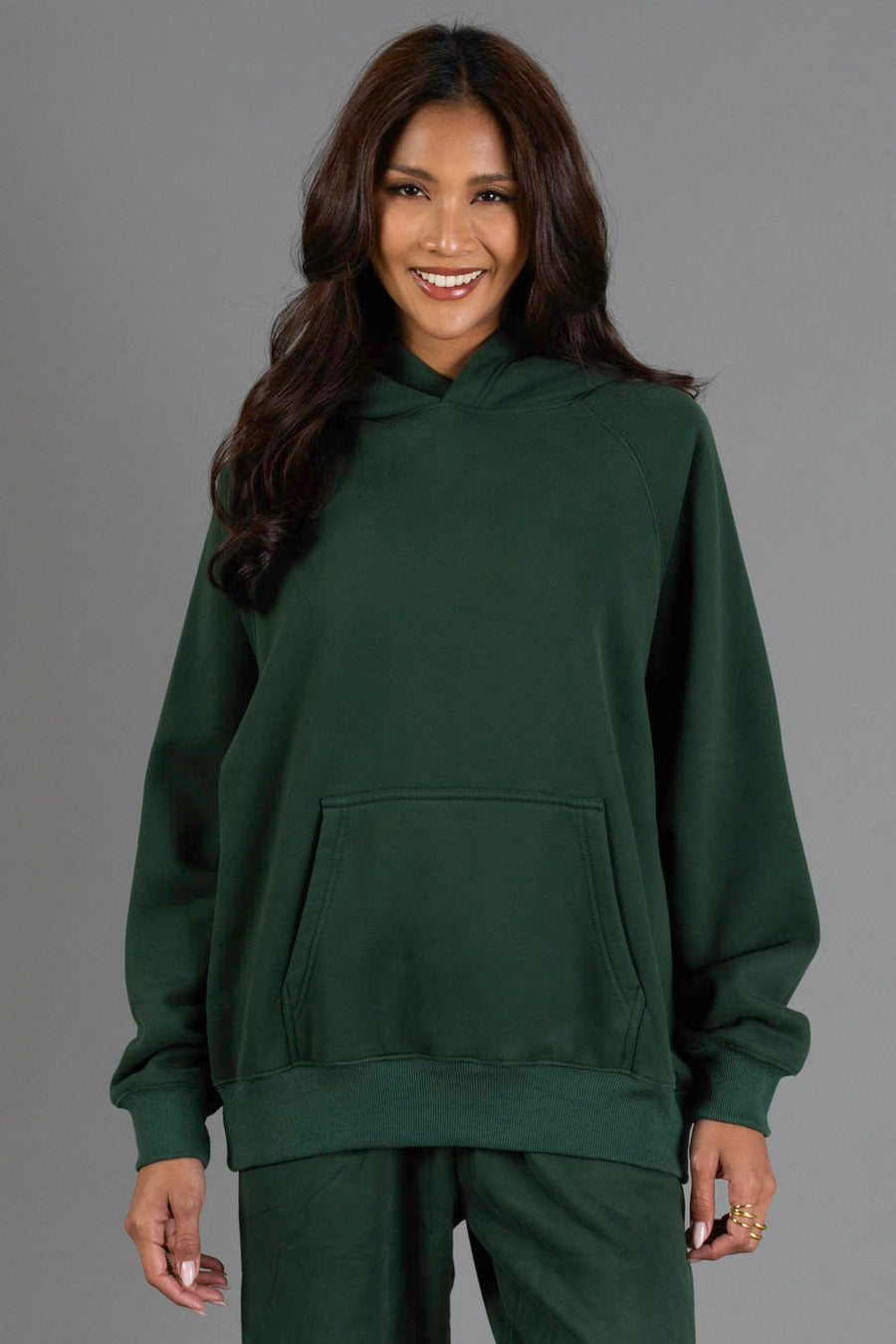 A woman with long dark hair smiles while wearing the Panache Apparel Collection’s Dark Green Panache Hooded Sweatshirt and matching pants, standing against a plain gray background.