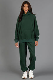 A woman stands and smiles at the camera against a plain gray background, wearing the Panache Apparel Collection’s Dark Green Panache Hooded Sweatshirt with matching sweatpants and white sneakers.