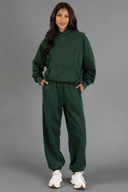 A person stands against a gray background, smiling with their hands in their pockets, wearing the Panache Apparel Collection’s Dark Green Panache Hooded Sweatshirt paired with matching sweatpants and white sneakers.