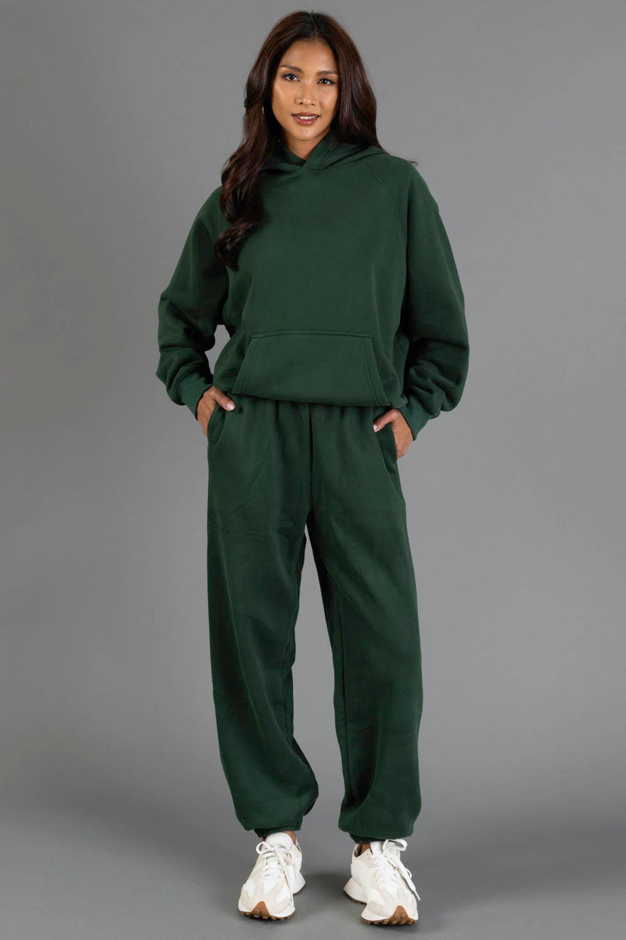 A person stands against a gray background, smiling with their hands in their pockets, wearing the Panache Apparel Collection’s Dark Green Panache Hooded Sweatshirt paired with matching sweatpants and white sneakers.