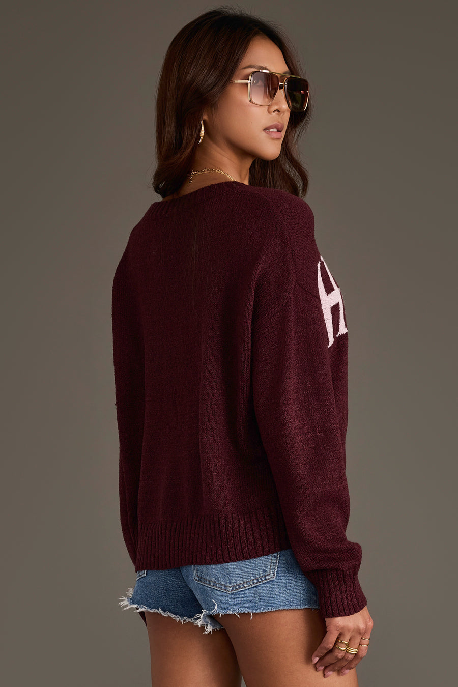 A woman stands sideways against a plain background, looking over her shoulder while wearing sunglasses, denim shorts, and the Maroon Howdy Lightweight Sweater from Panache Apparel Collection.