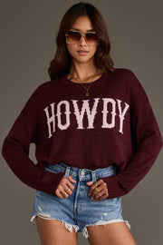 A woman stands confidently with her hands on her hips against a plain background, wearing the Panache Apparel Collection’s Maroon Howdy Lightweight Sweater, featuring bold white “HOWDY” lettering, paired with sunglasses and distressed denim shorts.