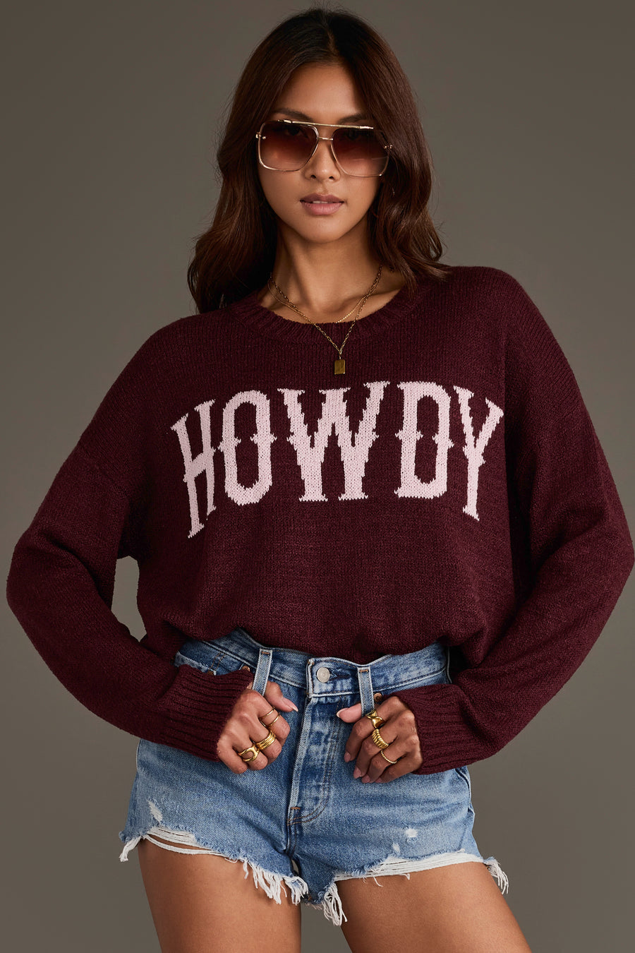 A woman stands confidently with her hands on her hips against a plain background, wearing the Panache Apparel Collection’s Maroon Howdy Lightweight Sweater, featuring bold white “HOWDY” lettering, paired with sunglasses and distressed denim shorts.