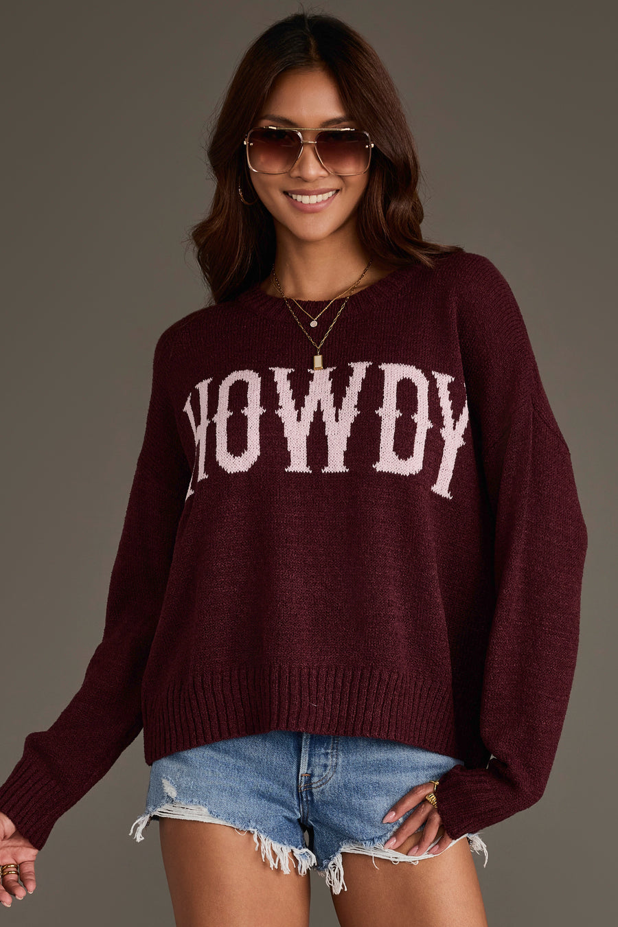 A woman wearing sunglasses and a Maroon Howdy Lightweight Sweater from Panache Apparel Collection, paired with denim shorts, is smiling and posing against a plain background.