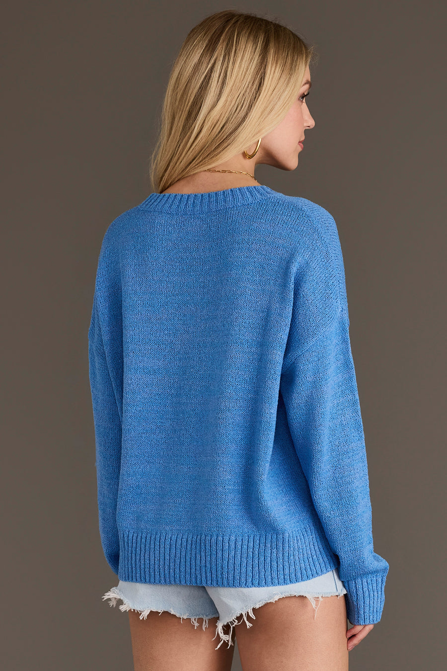 A woman with blonde hair faces away, wearing the Panache Apparel Collection Blue Howdy Lightweight Sweater and light denim cut-off shorts, standing against a plain brown background.