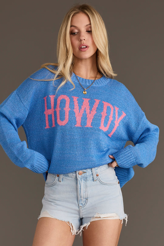A woman models the Panache Apparel Collection’s Blue Howdy Lightweight Sweater, featuring “HOWDY” in pink letters, paired with light denim shorts and posing against a plain background with one hand on her hip.