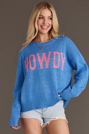 A smiling woman with long blonde hair wears the Panache Apparel Collection Blue Howdy Lightweight Sweater, featuring “HOWDY” in pink letters, paired with light denim shorts while standing against a plain background.