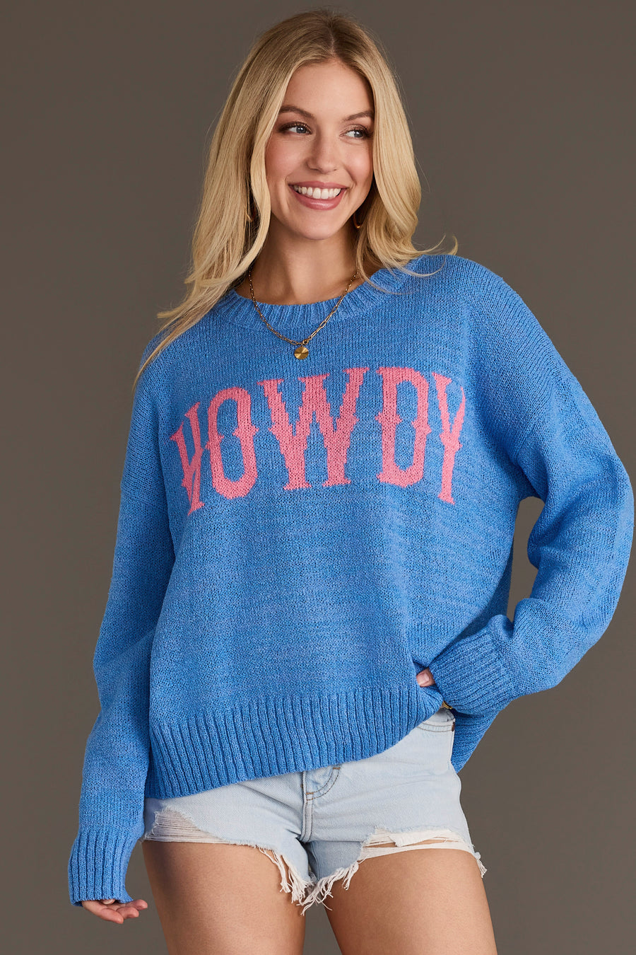 A smiling woman with long blonde hair wears the Panache Apparel Collection Blue Howdy Lightweight Sweater, featuring “HOWDY” in pink letters, paired with light denim shorts while standing against a plain background.