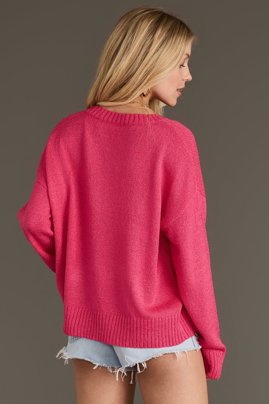 A person with long blonde hair, facing away from the camera against a plain brown background, wears the Pink Howdy Lightweight Sweater from Panache Apparel Collection paired with light blue denim shorts.