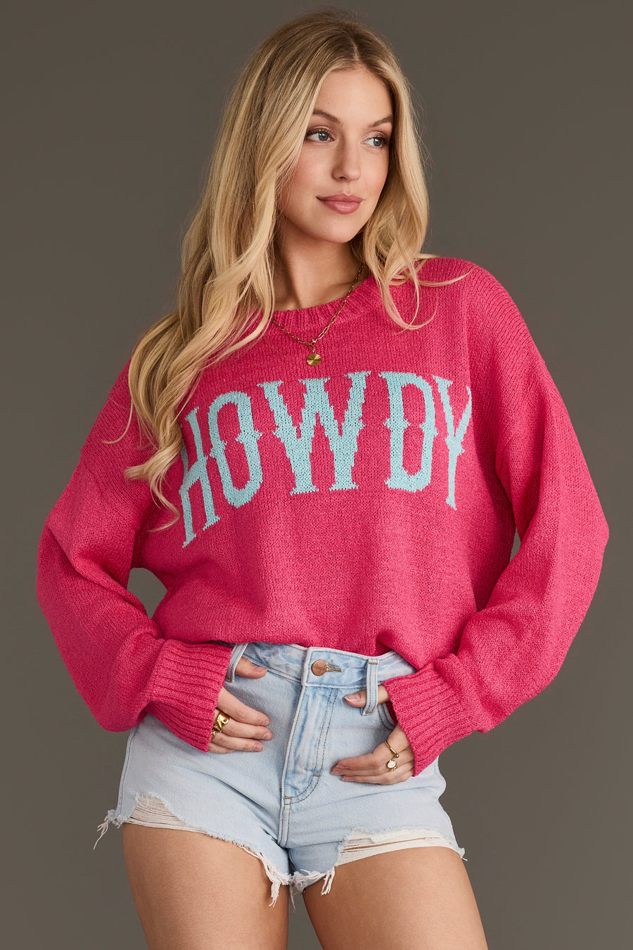 A person with long blonde hair wears the Panache Apparel Collection’s Pink Howdy Lightweight Sweater and light denim shorts, standing against a plain gray background.