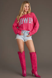 A woman poses against a brown background wearing the Pink Howdy Lightweight Sweater from Panache Apparel Collection, paired with light denim shorts and tall pink cowboy boots.