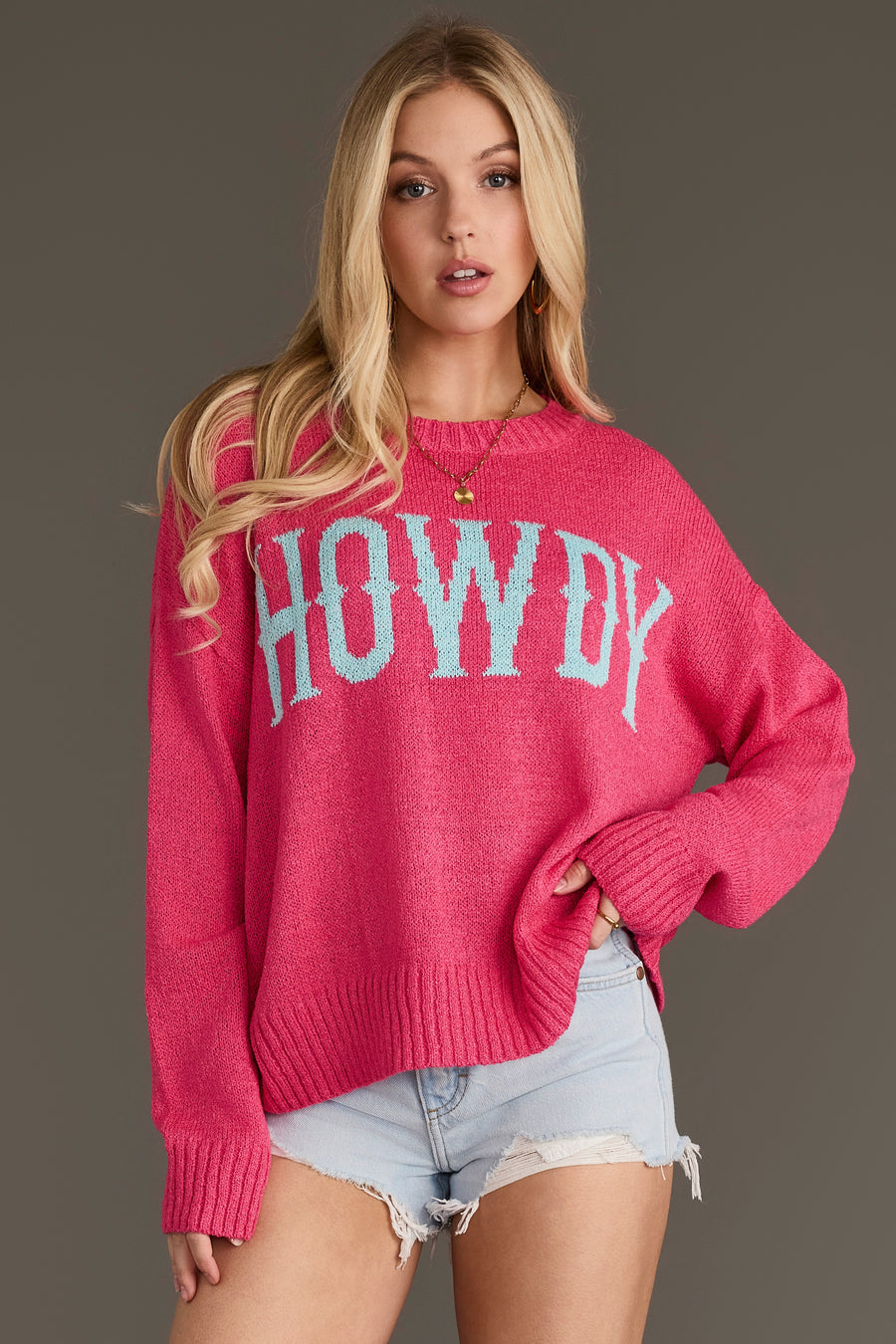 A woman models the Pink Howdy Lightweight Sweater from Panache Apparel Collection, featuring bold blue lettering across the front. She pairs it with light denim shorts, her long blonde hair cascading as she stands against a simple gray background.