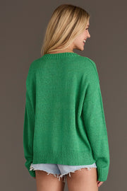 A woman with long blond hair, wearing the Green Howdy Lightweight Sweater from Panache Apparel Collection and frayed denim shorts, stands facing away and smiles against a plain brown background.