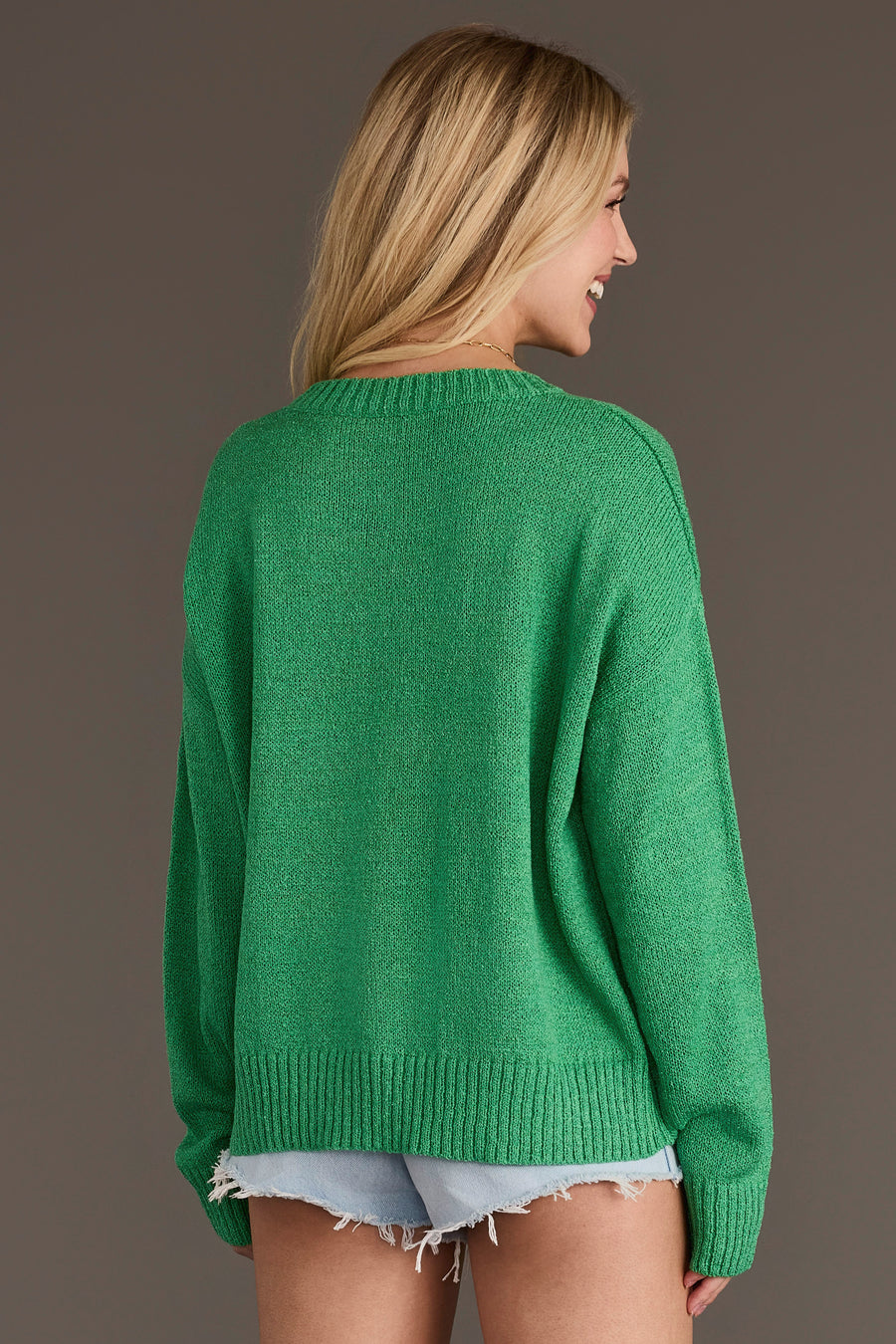 A woman with long blond hair, wearing the Green Howdy Lightweight Sweater from Panache Apparel Collection and frayed denim shorts, stands facing away and smiles against a plain brown background.