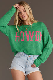 A woman stands against a plain gray background, looking down and touching her hair, wearing the Panache Apparel Collection Green Howdy Lightweight Sweater with pink lettering and light denim shorts.