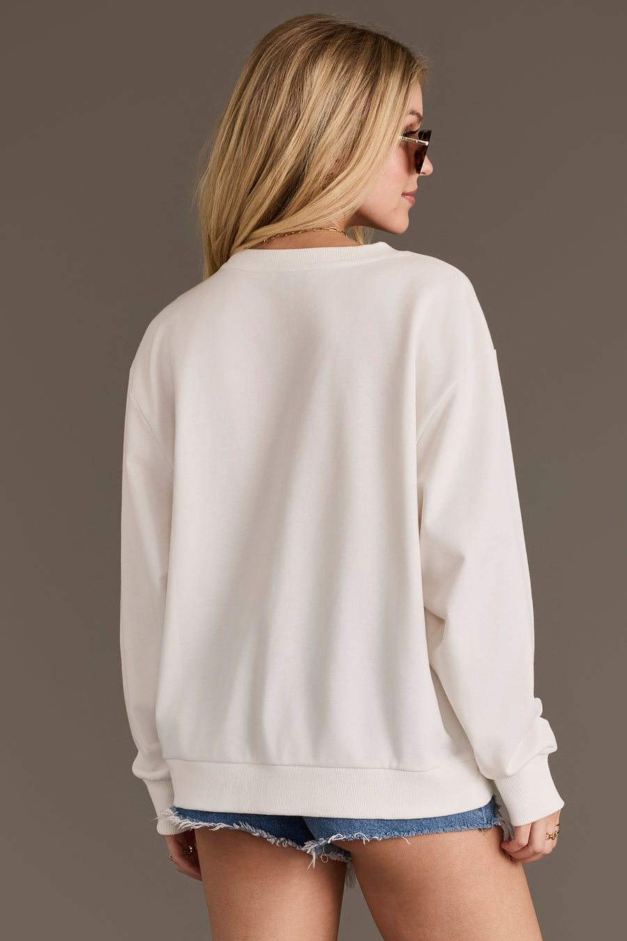 A woman with long blonde hair, wearing sunglasses, the Howdy Honey Sweatshirt by Panache Apparel Collection, and denim shorts stands facing away from the camera against a plain background.
