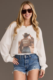 A woman wearing sunglasses, denim shorts, and the Panache Apparel Collection’s Howdy Honey Sweatshirt stands smiling with one hand on her hip against a plain background.