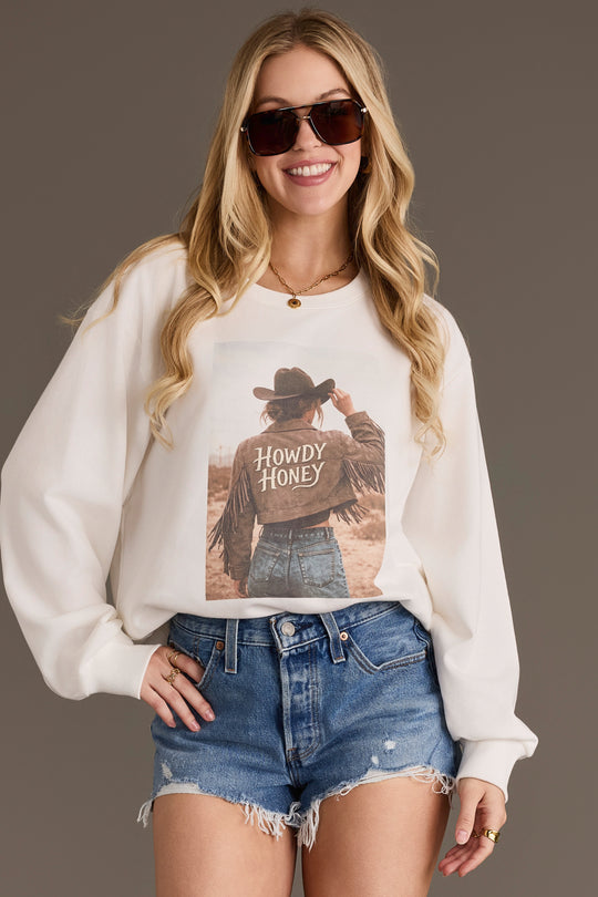 A woman wearing sunglasses, denim shorts, and the Panache Apparel Collection’s Howdy Honey Sweatshirt stands smiling with one hand on her hip against a plain background.