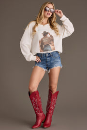A woman stands confidently against a plain background, wearing the Panache Apparel Collection’s Howdy Honey Sweatshirt styled with denim shorts, red cowboy boots, and sunglasses, one hand on her hip, the other adjusting her shades.