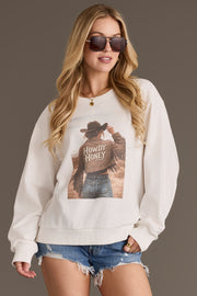 A woman with long, wavy blonde hair wears the Panache Apparel Collection Howdy Honey Sweatshirt, featuring a graphic of a person in a cowboy hat and jacket with the words “Howdy Honey,” paired with denim shorts and sunglasses.