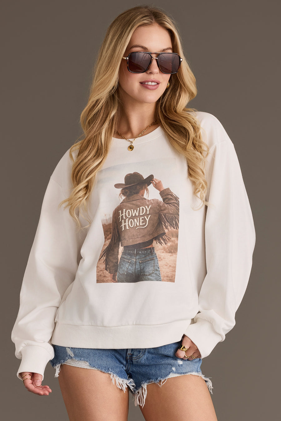 A woman with long, wavy blonde hair wears the Panache Apparel Collection Howdy Honey Sweatshirt, featuring a graphic of a person in a cowboy hat and jacket with the words “Howdy Honey,” paired with denim shorts and sunglasses.
