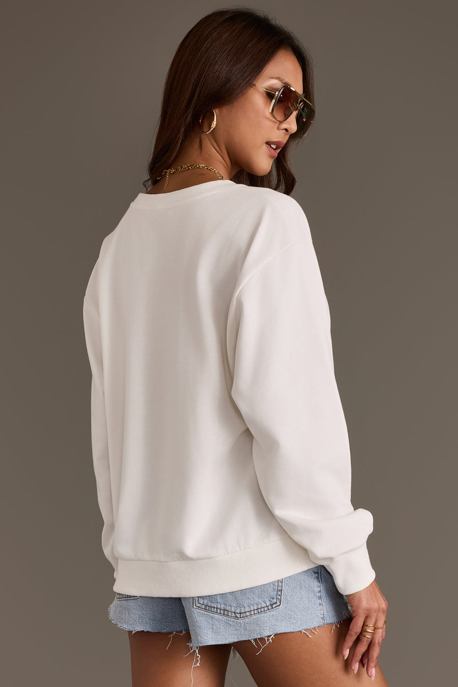 A woman wearing sunglasses, ripped denim shorts, and the Panache Apparel Collection Western Sign Sweatshirt is standing with her back to the camera against a plain, neutral background.