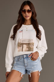 A woman wears the Panache Apparel Collection Western Sign Sweatshirt, featuring a Howdy Honey graphic and American flag, paired with light blue distressed denim shorts and sunglasses, standing against a plain background with one hand in her pocket.