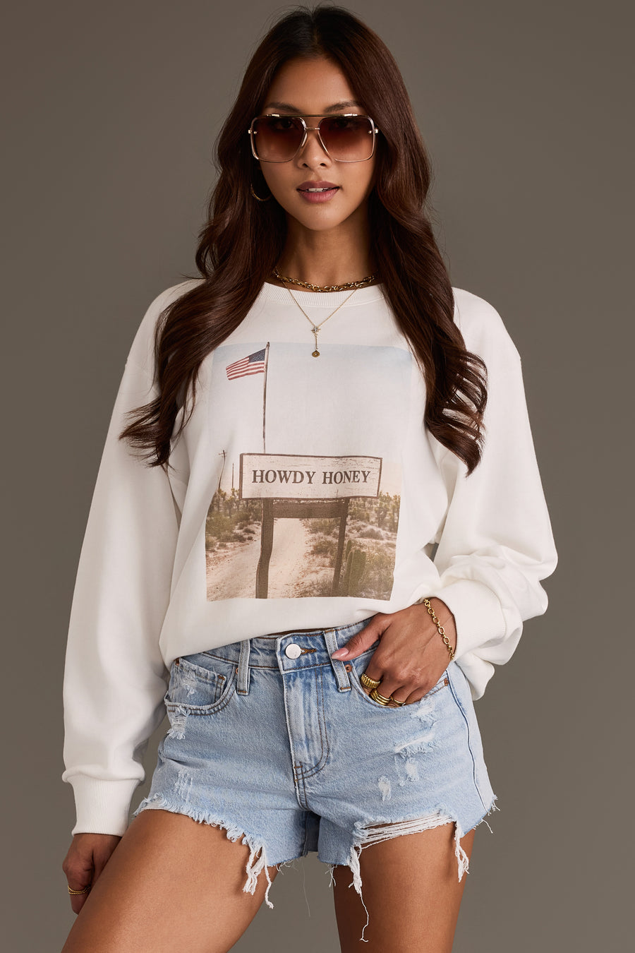 A woman wears the Panache Apparel Collection Western Sign Sweatshirt, featuring a Howdy Honey graphic and American flag, paired with light blue distressed denim shorts and sunglasses, standing against a plain background with one hand in her pocket.