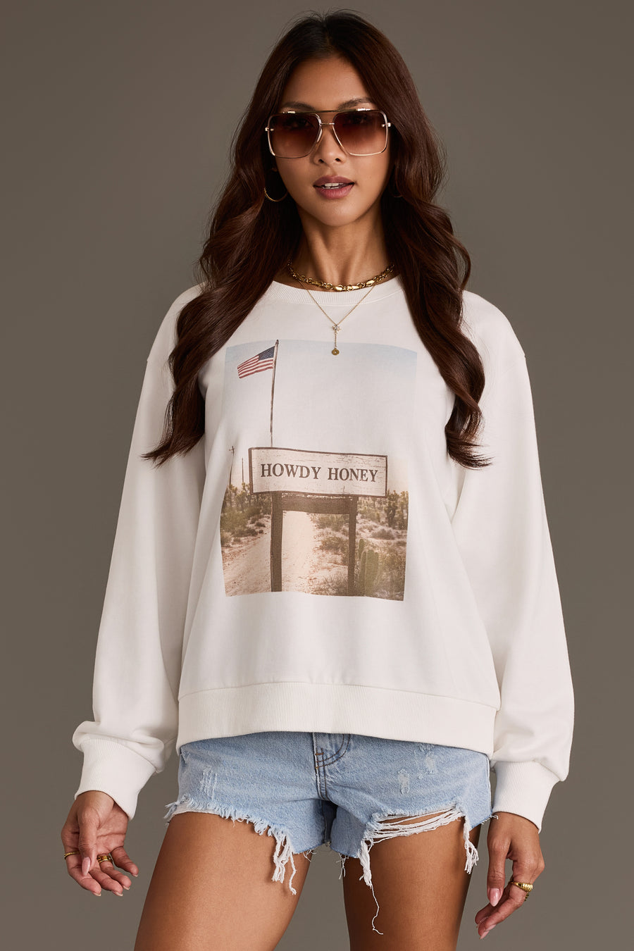A woman wearing sunglasses, gold jewelry, denim shorts, and the Panache Apparel Collection Western Sign Sweatshirt, featuring a “Howdy Honey” graphic, stands against a plain gray background.