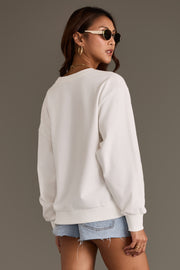 A woman wearing sunglasses, hoop earrings, and denim shorts models the Panache Apparel Collection’s Patriotic Cowboy Sweatshirt, standing with her back turned and glancing over her shoulder against a plain background.