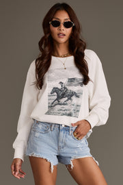 A woman stands against a plain background wearing sunglasses, a gold necklace, light blue denim shorts, and the Panache Apparel Collection’s Patriotic Cowboy Sweatshirt, a white top featuring a horse and American flag print.