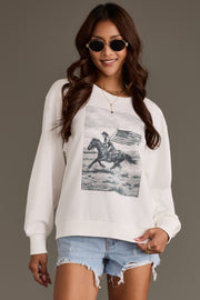 A woman poses and smiles against a neutral background, wearing the Panache Apparel Collection’s Patriotic Cowboy Sweatshirt, featuring a print of a cowboy on horseback holding an American flag, styled with sunglasses and distressed denim shorts.