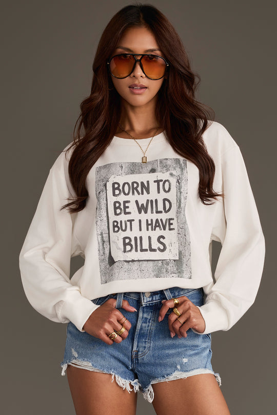 A woman stands with her hands on her hips against a neutral background, wearing sunglasses, distressed denim shorts, and the Panache Apparel Collection’s “Born to Be Wild Sweatshirt,” featuring the playful phrase “BORN TO BE WILD BUT I HAVE BILLS.”.
