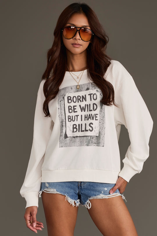 A woman wearing sunglasses and denim shorts stands against a plain background with one hand in her pocket, styling the Panache Apparel Collection “Born to Be Wild Sweatshirt,” featuring the playful slogan “BORN TO BE WILD BUT I HAVE BILLS” printed on the front.