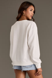 A woman with long brown hair wears the Panache Apparel Collection’s Coyote Saloon Sweatshirt in white paired with light blue denim shorts. She faces away, looking over her shoulder against a plain brown background.