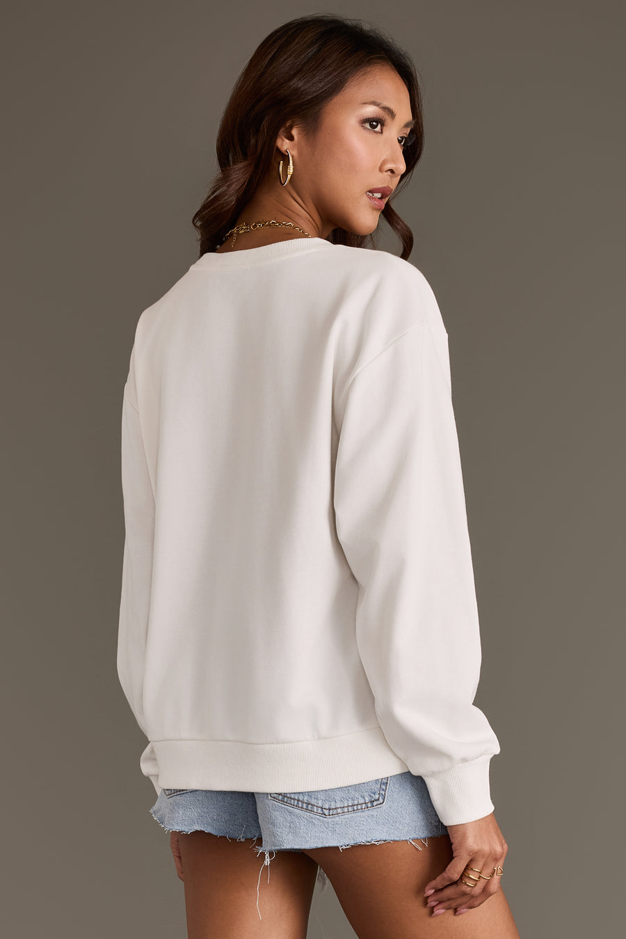 A woman with long brown hair wears the Panache Apparel Collection’s Coyote Saloon Sweatshirt in white paired with light blue denim shorts. She faces away, looking over her shoulder against a plain brown background.