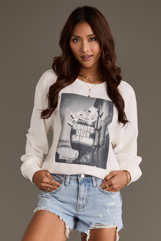 A woman wearing the Panache Apparel Collection Coyote Saloon Sweatshirt in white and light blue denim shorts poses against a plain gray background. She has long, wavy brown hair and accessorizes with gold jewelry.