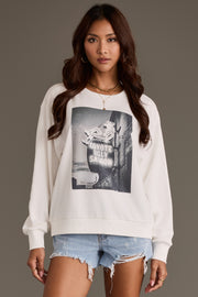 A woman with long brown hair wears the Panache Apparel Collection “Coyote Saloon Sweatshirt,” featuring a sign graphic, paired with light denim shorts, while standing against a plain gray background.