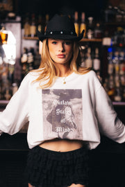A woman wearing the Panache Apparel Collection’s Outlaw State of Mind Sweatshirt and a black cowboy hat stands in front of a bar lined with bottles, embodying bold, Western-inspired style.