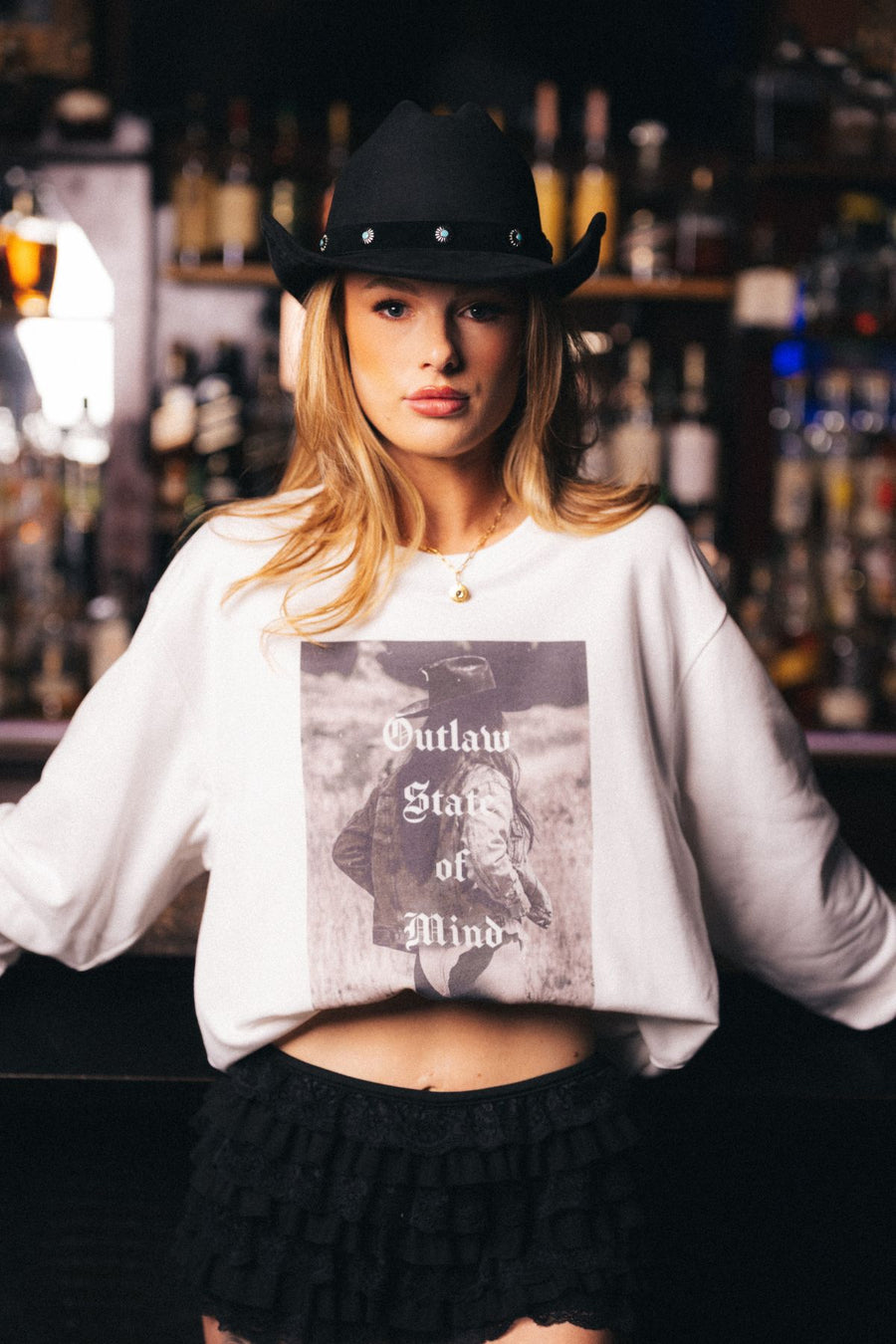 A woman wearing the Panache Apparel Collection’s Outlaw State of Mind Sweatshirt and a black cowboy hat stands in front of a bar lined with bottles, embodying bold, Western-inspired style.